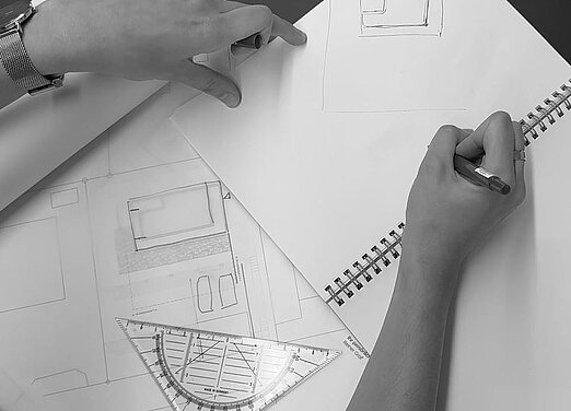 Ein Skizzenbuch mit Entwürfen für ein zukünftiges Architektenhaus sowie ein Geodreieck liegen auf einem Tisch.