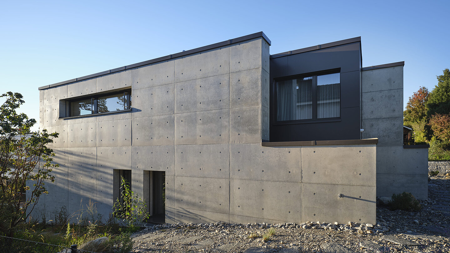 A view of the side wall of the Akinori detached house. The building stands out from the otherwise green landscape with its concrete look.
